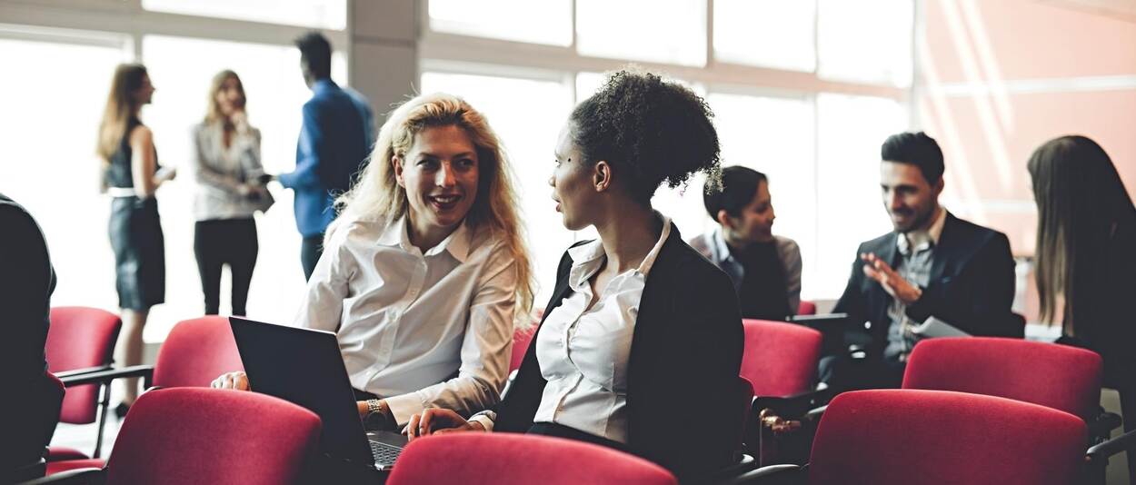 A view of participants in a conference room: some people are still sitting and talking together, some are standing around the room