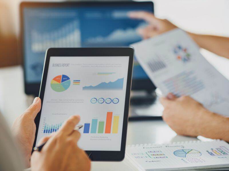 A hand holding an iPad with graphs on the screen, next to a paper with graphs and in the background a computer screen with graphs (out of focus)